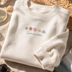 May include: Cream-colored sweatshirt with embroidered seashells and the word "Summertime" in blue script. The design includes a starfish, coral, a scallop shell, a spiral shell, and a wave. The sweatshirt has a crew neck and ribbed cuffs.
