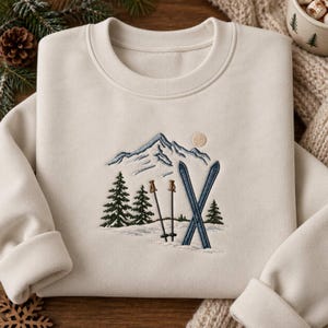 May include: A cream-colored crewneck sweatshirt featuring an embroidered design of a snow-capped mountain range, a pale sun, evergreen trees, two brown ski poles, and a pair of blue skis positioned in a snowy landscape on the front center.