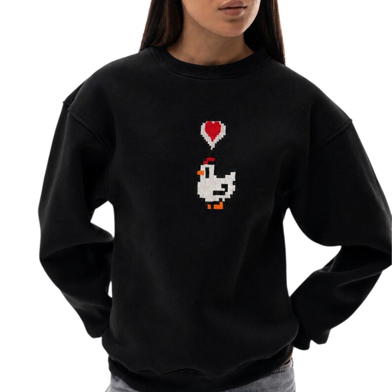May include: Black sweatshirt featuring a pixelated design of a white chicken with a red comb and a red heart above it. The sweatshirt has long sleeves and a crew neck.
