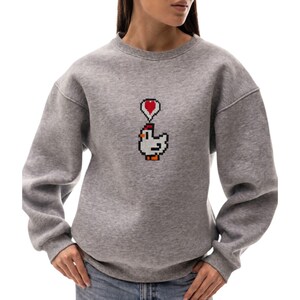 May include: A heather gray sweatshirt featuring a pixelated chicken graphic with a red heart above its head. The long-sleeved crewneck top is made of a soft material and is suitable for casual wear.