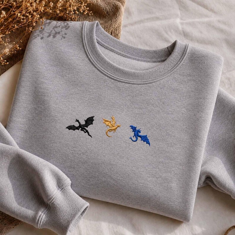 Op de afbeelding: Een grijs sweatshirt met geborduurde draken in zwart, goud en blauw. De crew neck sweatshirt heeft lange mouwen en een relaxte pasvorm. De draken zijn gestileerd en lijken te vliegen. De sweatshirt is gemaakt van een zacht materiaal.