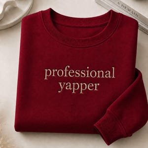 May include: A deep red crewneck sweatshirt featuring the embroidered text "professional yapper" in a light beige, sans-serif font centred on the chest. The garment has long sleeves with ribbed cuffs and a matching ribbed neckline.