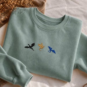 Op de afbeelding: Een lichtgroene sweatshirt met geborduurde draken in zwart, goud en blauw. De sweatshirt met ronde hals heeft lange mouwen en een relaxte pasvorm. De draken zijn horizontaal over de borst geplaatst.