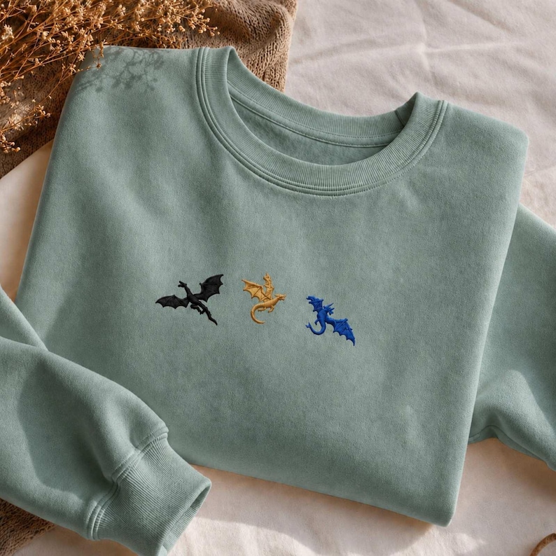 Op de afbeelding: Een lichtgroene sweatshirt met geborduurde draken in zwart, goud en blauw. De sweatshirt met ronde hals heeft lange mouwen en een relaxte pasvorm. De draken zijn horizontaal over de borst geplaatst.