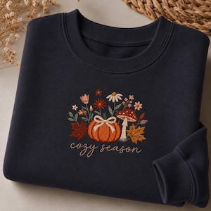 Op de afbeelding: Een donkerblauw sweatshirt met ronde hals en een geborduurd ontwerp van een oranje pompoen met een witte strik, een rood-witte paddenstoel, herfstbladeren en diverse bloemen, met de tekst "cozy season" gestikt in lichtbruin garen.