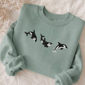 Puede incluir: Una sudadera verde salvia con tres orcas bordadas en blanco y negro. Las orcas están dispuestas en la parte delantera de la sudadera. La prenda está hecha de un material suave y es adecuada para ropa casual.