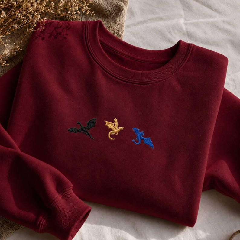 Op de afbeelding: Een bordeauxrode sweatshirt met geborduurde draken in zwart, goud en blauw. De sweatshirt met ronde hals heeft lange mouwen en een zacht uiterlijk. De draken zijn in een rij over de borst geborduurd.