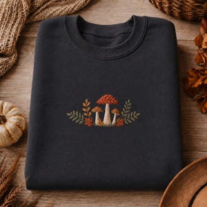 Op de afbeelding: Een donkergrijs sweatshirt met ronde hals en een centrale borduursel van drie paddenstoelen in rood- en bruintinten, omringd door kleine groene blaadjes en oranje herfstbladeren in een gedetailleerd botanisch ontwerp.