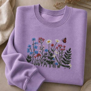 May include: A lavender sweatshirt with an embroidered floral design. The embroidery features a variety of colorful flowers, including blue, red, and white blooms, along with a small butterfly. The sweatshirt has a crew neck and long sleeves.