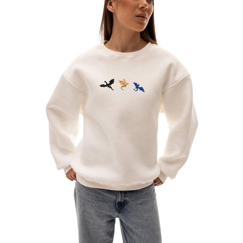 Op de afbeelding: Cr&egrave;mekleurige sweatshirt met drie geborduurde draken silhouetten in zwart, goud en blauw. De crew neck sweatshirt heeft lange mouwen en is gemaakt van een zacht materiaal. De draken zijn horizontaal over de borst geplaatst.