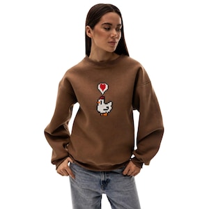 May include: Brown sweatshirt featuring a pixelated chicken design with a red heart above it. The sweatshirt has a crew neck and long sleeves. The model is wearing blue jeans.