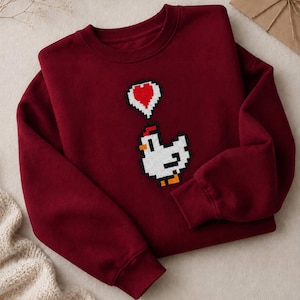 May include: Burgundy sweatshirt featuring a pixelated white chicken with a red comb and orange feet. A pixelated red heart floats above the chicken. The sweatshirt has a crew neck and long sleeves.