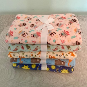 May include: A stack of five folded fabric pieces tied with a white ribbon. The top fabric is light pink with cowboy cats, the second is light green with boots, the third is cream with foxes, the fourth is blue with bears, and the bottom is dark blue with suns.