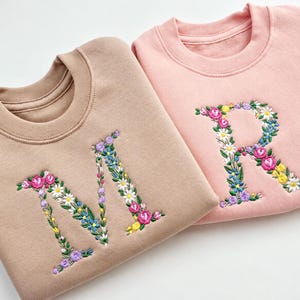May include: A tan sweatshirt featuring an embroidered letter M and a pink sweatshirt featuring an embroidered letter R. Both letters are decorated with colourful floral patterns including pink, yellow, white, and blue flowers with green leaves.