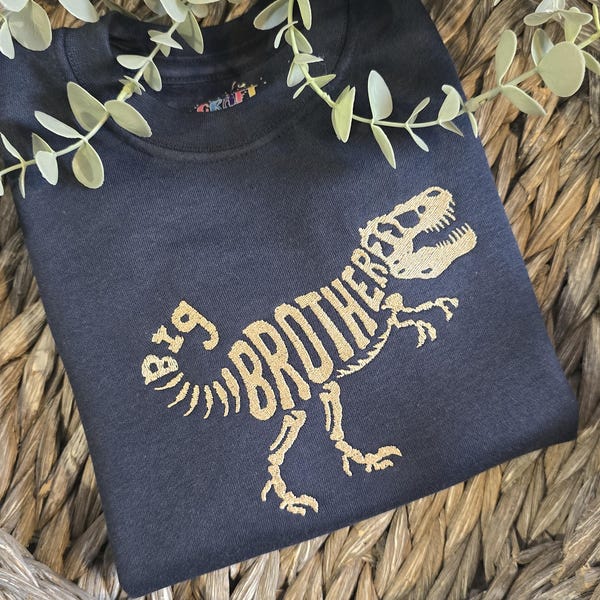 Puede incluir: Una sudadera negra de cuello redondo con un gráfico dorado de un esqueleto de dinosaurio. El texto "BIG BROTHER" está integrado en la estructura ósea del dinosaurio, donde "BIG" forma la cola y "BROTHER" forma el cuerpo del animal.