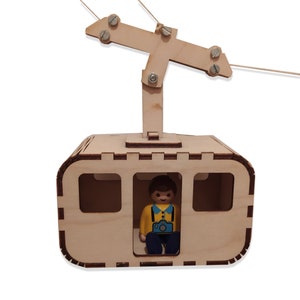 May include: A wooden toy cable car with a small figure of a person inside. The cable car is brown and has a window on each side. The figure is wearing a yellow shirt, blue trousers, and a blue hat. The cable car is suspended from a wire.