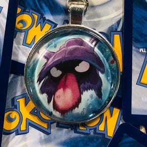 May include: A silver charm with a purple and black Pokemon character with a large red nose. The character is surrounded by a blue and white background.