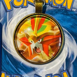 Könnte beinhalten: Ein bronzenfarbener Anhänger mit einem Nahaufnahmebild eines Pokémon-Charakters in Rot, Gelb und Grün. Der Charakter hat einen grimmigen Ausdruck mit scharfen Zähnen und einer sternförmigen Markierung auf der Stirn. Der Anhänger ist von einer klaren Glaskuppel umgeben.