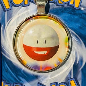 Può includere: Un ciondolo d'argento con una faccia rotonda in vetro trasparente che presenta un'immagine di un personaggio dei cartoni animati Pokémon rosso e bianco con un viso sorridente. Il personaggio ha due occhi neri e uno sfondo bianco.