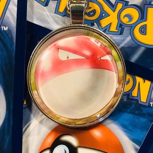 Può includere: Un ciondolo a forma di charm in argento con un cabochon di vetro rotondo che raffigura un personaggio di cartone animato rosso e bianco di Pokemon con un'espressione minacciosa.