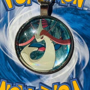 Könnte beinhalten: Eine silberne Anhänger-Kette mit einem runden Glascabochon, der ein farbenfrohes Bild eines Pokemon-Charakters zeigt. Der Charakter ist eine weiße und rosafarbene Seeschlange vor blauem Hintergrund.