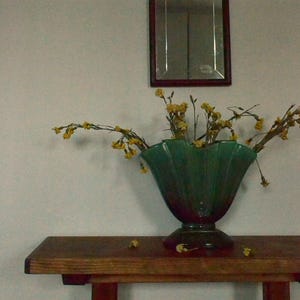 Puede incluir: Un arreglo decorativo con un jarrón verde oscuro con flores amarillas sobre una mesa de madera. Un pequeño espejo rectangular con un marco marrón oscuro cuelga de la pared sobre la mesa. La escena se desarrolla contra una pared clara.
