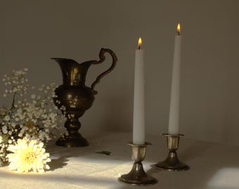Può includere: Due candele bianche con fiamme che bruciano in candelabri d'argento su una tovaglia bianca con un'anfora di ottone e fiori bianchi sullo sfondo.