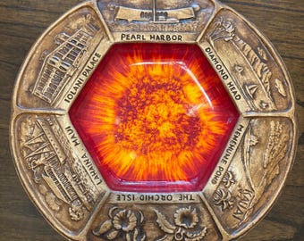 Könnte beinhalten: Ein rundes dekoratives Metalltablett mit einem zentralen hexagonalen rot-orangen Sonnenstrahlenmuster. Der Rand zeigt geprägte hawaiianische Wahrzeichen, darunter U.S.S. Arizona, Pearl Harbor, Iolani Palace, Diamond Head, Lahaina, Maui, Menemene Pond, Kauai und Hawaii.