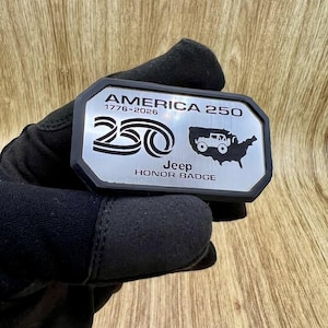 May include: A rectangular metal badge with a black border and a silver face. The text reads AMERICA 250, 1776-2026, Jeep, and HONOR BADGE. It features a graphic of the number 250 and a silhouette of the United States containing a small Jeep vehicle.