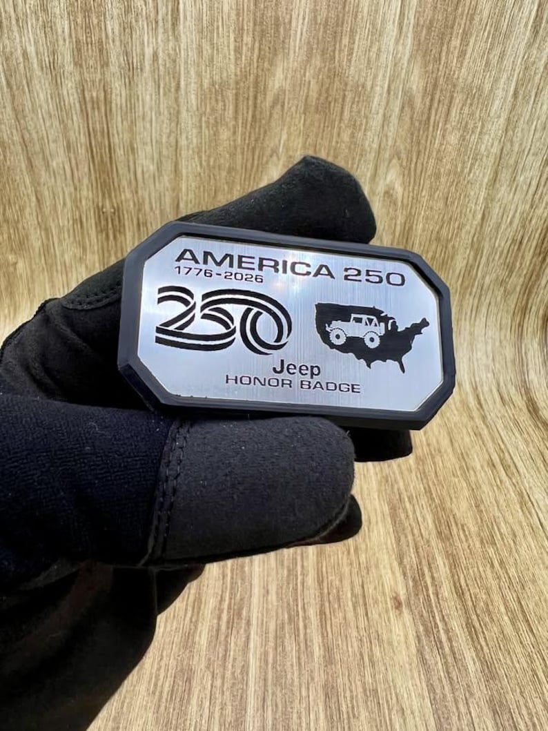 May include: A rectangular metal badge with a black border and a silver face. The text reads AMERICA 250, 1776-2026, Jeep, and HONOR BADGE. It features a graphic of the number 250 and a silhouette of the United States containing a small Jeep vehicle.