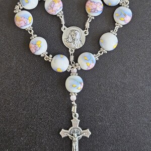 May include: A rosary with white, floral-patterned beads and silver-toned metal accents. The rosary features a crucifix with a figure of Jesus and a medallion with a religious figure.