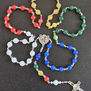 May include: A collection of rosaries featuring colorful beads in red, yellow, green, white, and blue. Each rosary is strung on a silver chain, with a cross pendant and a decorative centerpiece. The beads are round and smooth.