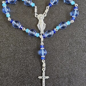 May include: A rosary with blue glass beads and silver-tone metal accents. The beads vary in size and shade, with a mix of light and dark blue. The centerpiece features a depiction of the Virgin Mary, and the cross has a detailed crucifix.