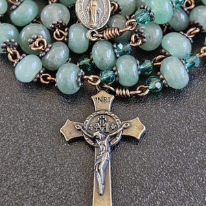 K&ouml;nnte beinhalten: Ein Rosenkranz mit hellgr&uuml;nen Perlen und einem Metallkruzifix. Die Perlen haben eine sanfte gr&uuml;ne Farbe, und die Metallkomponenten haben ein antikes Finish. Das Kruzifix zeigt eine detaillierte Darstellung von Jesus Christus.