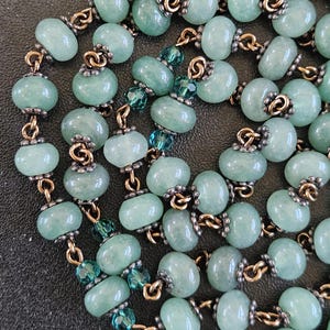 K&ouml;nnte beinhalten: Halskette mit hellgr&uuml;nen, ovalen Perlen, die durch goldfarbene Metallglieder verbunden sind. Das Design umfasst auch kleinere, facettierte teal-farbene Perlen. Die Perlen haben ein leicht durchscheinendes Aussehen.