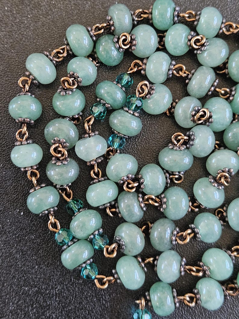 K&ouml;nnte beinhalten: Halskette mit hellgr&uuml;nen, ovalen Perlen, die durch goldfarbene Metallglieder verbunden sind. Das Design umfasst auch kleinere, facettierte teal-farbene Perlen. Die Perlen haben ein leicht durchscheinendes Aussehen.