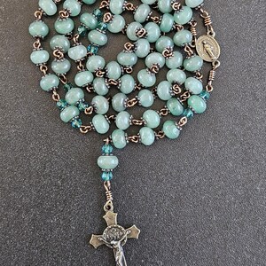 K&ouml;nnte beinhalten: Ein Rosenkranz mit hellgr&uuml;nen, ovalen Perlen an einer bronzefarbenen Kette. Er hat einen Kreuzanh&auml;nger und eine Medaille mit einer religi&ouml;sen Figur. Die Perlen sind mit kleineren, blau-gr&uuml;nen Perlen durchsetzt.