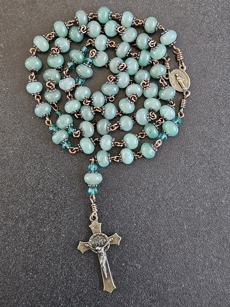 K&ouml;nnte beinhalten: Ein Rosenkranz mit hellgr&uuml;nen, ovalen Perlen an einer bronzefarbenen Kette. Er hat einen Kreuzanh&auml;nger und eine Medaille mit einer religi&ouml;sen Figur. Die Perlen sind mit kleineren, blau-gr&uuml;nen Perlen durchsetzt.