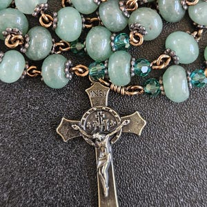 K&ouml;nnte beinhalten: Ein Rosenkranz mit hellgr&uuml;nen Edelsteinperlen und einem detaillierten Bronzekreuz. Die Perlen sind mit goldfarbenem Draht und kleineren gr&uuml;nen Kristallperlen verbunden. Das Kruzifix tr&auml;gt oben die Buchstaben INRI.
