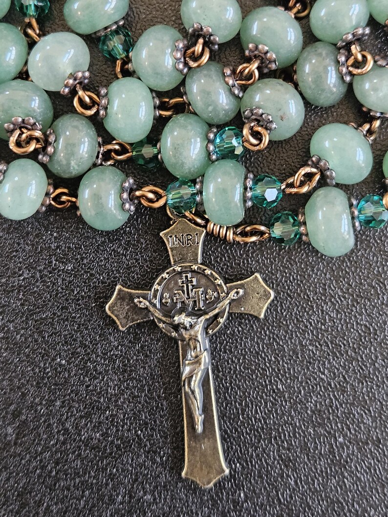 K&ouml;nnte beinhalten: Ein Rosenkranz mit hellgr&uuml;nen Edelsteinperlen und einem detaillierten Bronzekreuz. Die Perlen sind mit goldfarbenem Draht und kleineren gr&uuml;nen Kristallperlen verbunden. Das Kruzifix tr&auml;gt oben die Buchstaben INRI.