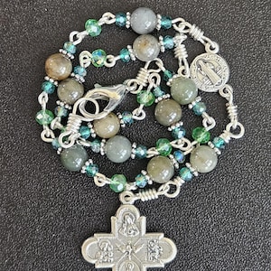 May include: A rosary with gray and green beads, silver-toned chain links, and a silver cross pendant. The cross features religious figures and symbols. The beads are a mix of round and faceted shapes. A small medallion is also attached.