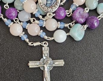 May include: A rosary featuring round beads in shades of purple, pale pink, and light blue, accented with small silver-toned spacers. The centerpiece is an oval medal depicting a figure holding a child, and the crucifix is a silver-toned metal cross.