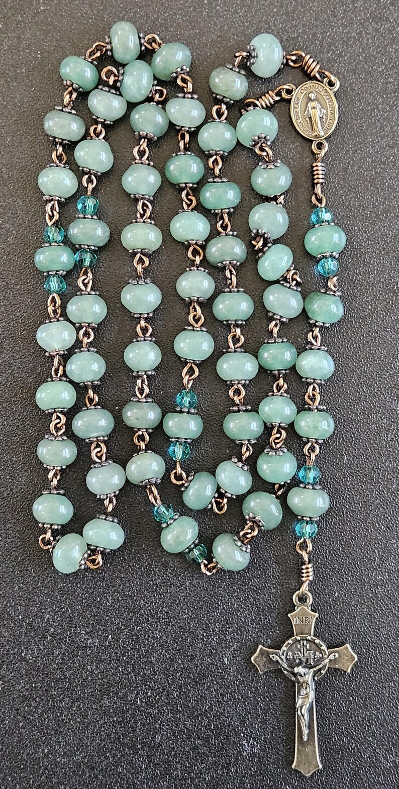 K&ouml;nnte beinhalten: Ein Rosenkranz mit hellgr&uuml;nen, ovalen Perlen und kupferfarbenen Metallgliedern. Der Rosenkranz hat einen Kreuzanh&auml;nger und eine kleine Medaille. Kleine blaue Perlen sind im gesamten Design verstreut.