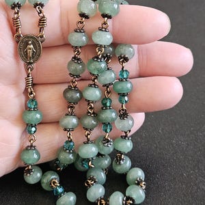 K&ouml;nnte beinhalten: Ein Rosenkranz mit hellgr&uuml;nen Jadeperlen und bronzefarbenen Metallakzenten. Die Perlen sind durch bronzefarbenen Draht verbunden und mit kleineren, teal-farbenen Perlen durchsetzt. Eine bronzefarbene Medaille mit einer religi&ouml;sen Figur ist sichtbar.