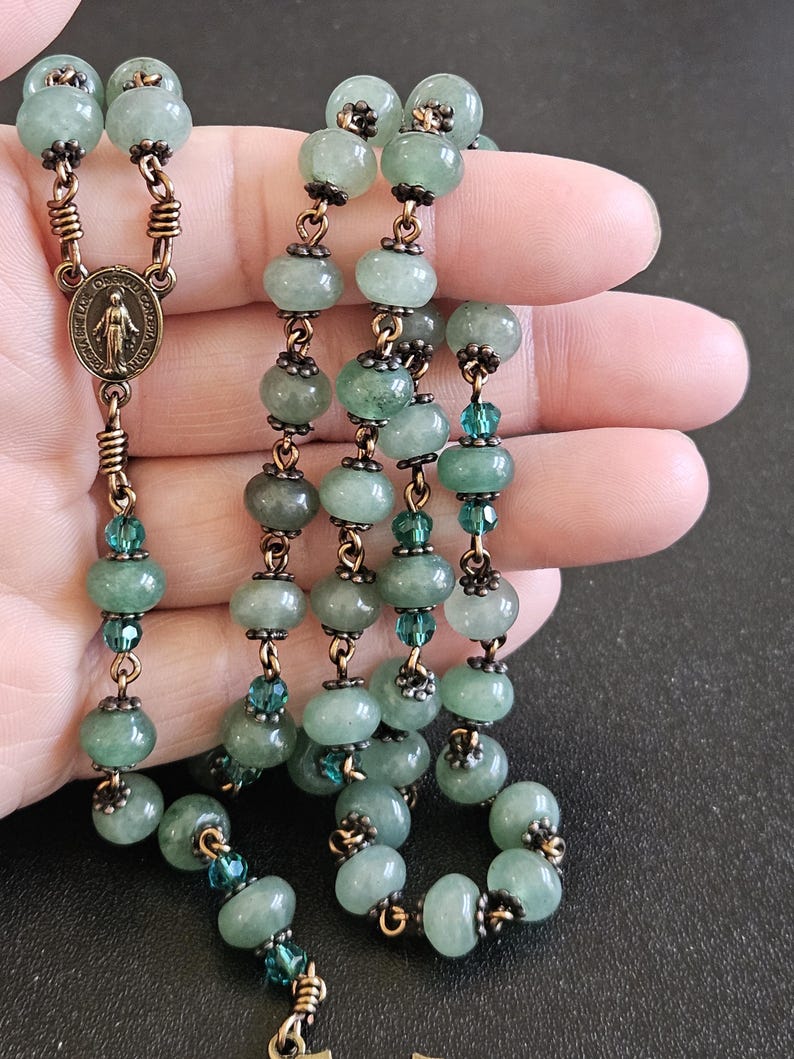K&ouml;nnte beinhalten: Ein Rosenkranz mit hellgr&uuml;nen Jadeperlen und bronzefarbenen Metallakzenten. Die Perlen sind durch bronzefarbenen Draht verbunden und mit kleineren, teal-farbenen Perlen durchsetzt. Eine bronzefarbene Medaille mit einer religi&ouml;sen Figur ist sichtbar.
