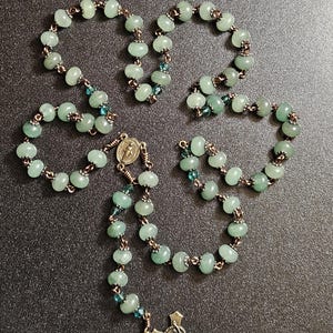 K&ouml;nnte beinhalten: Ein Rosenkranz mit hellgr&uuml;nen Perlen und einem silberfarbenen Kruzifix. Die Perlen sind durch kupferfarbenen Draht verbunden. Kleine blaue Perlen sind im Rosenkranz verstreut.