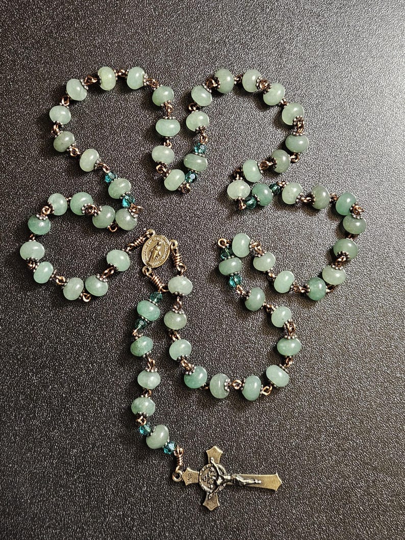 K&ouml;nnte beinhalten: Ein Rosenkranz mit hellgr&uuml;nen Perlen und einem silberfarbenen Kruzifix. Die Perlen sind durch kupferfarbenen Draht verbunden. Kleine blaue Perlen sind im Rosenkranz verstreut.