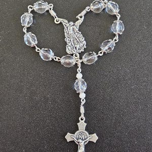 May include: A silver rosary with clear, faceted beads. The rosary features a detailed silver centerpiece depicting an angel and a crucifix with the figure of Jesus. The beads are connected by silver wire.
