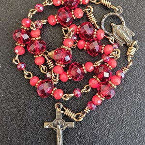 May include: A rosary with faceted red beads and smaller red beads strung on a gold-colored chain. The rosary features a crucifix and a small pendant with a religious figure.