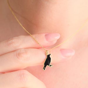 Op de afbeelding: Een gouden ketting met een zwarte vogelhanger. De vogel is gesilhouetteerd tegen een gouden achtergrond.